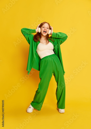 Cheerful young girl, teenager with red hair wearing headphones listening songs and smiling over yellow studio background. Music lover photo