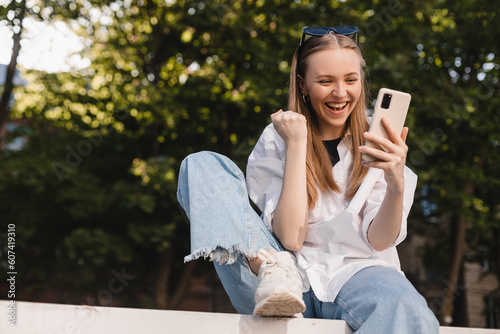 Smiling young girl sitting on ground browsing internet with cellphone gesturing like winner. Excited face expression, unable to believe that she bet played, win in lottery, can't believe in fortune.