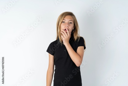 Nervous puzzled beautiful teen girl wearing black dress over white studio background opens mouth from surprise, reacts on sudden news.