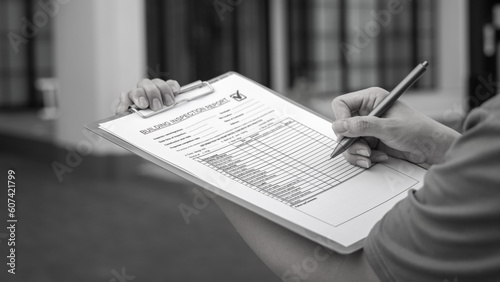 An engineer hand is using a pen checking on the building inspection report form to QC building quality of the house (background). Export industrial working scene concept. Close-up and selective focus.