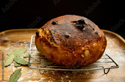 Fresh sourdough raisin bread loaf on a cooling rack on a wooden tray with bay leaves and flour against a black background photo