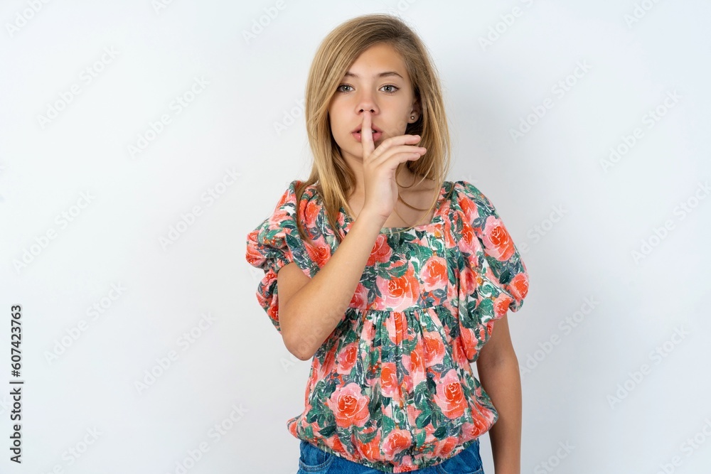 beautiful teen girl wearing flowered dress over white studio background makes silence gesture, keeps finger over lips. Silence and secret concept. beautiful teen girl wearing flowered dress over white studio background makes silence gesture, keeps finger over lips. Silence and secret concept.