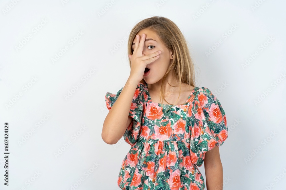 beautiful teen girl wearing flowered dress over white studio background peeking in shock covering face and eyes with hand, looking through fingers with embarrassed expression. beautiful teen girl wearing flowered dress over white studio background peeking in shock covering face and eyes with hand, looking through fingers with embarrassed expression.