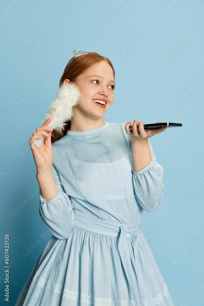 Queen, beautiful princess with red hair holding hand fan and recording audio message in messenger over blue studio background. Concept of social networks, gadgets Queen, beautiful princess with red hair holding hand fan and recording audio message in messenger over blue studio background. Concept of social networks, gadgets