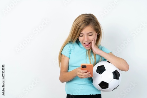 beautiful caucasian teen girl wearing sportswear holding a football ball over white wall holding in hands cell reading sms using new app 5g © Jihan