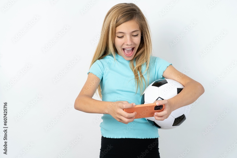 Portrait of an excited beautiful caucasian teen girl wearing sportswear holding a football ball over white wall playing games on mobile phone. Portrait of an excited beautiful caucasian teen girl wearing sportswear holding a football ball over white wall playing games on mobile phone.