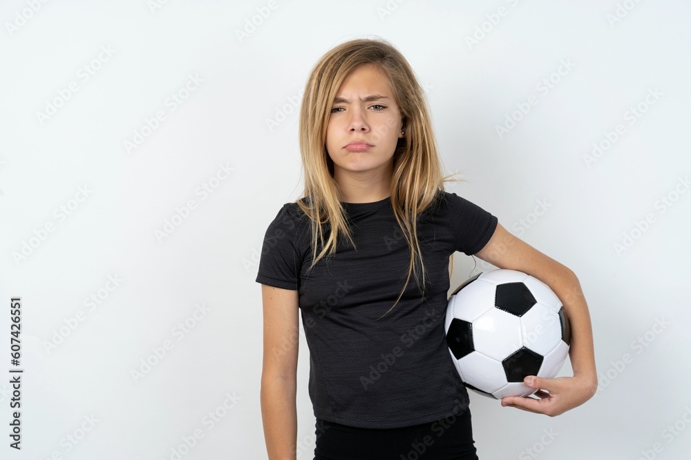 Offended dissatisfied beautiful caucasian teen girl wearing sportswear holding a football ball over white wall with moody displeased expression at camera being disappointed by something