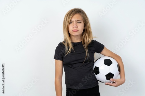 Offended dissatisfied beautiful caucasian teen girl wearing sportswear holding a football ball over white wall with moody displeased expression at camera being disappointed by something © Jihan