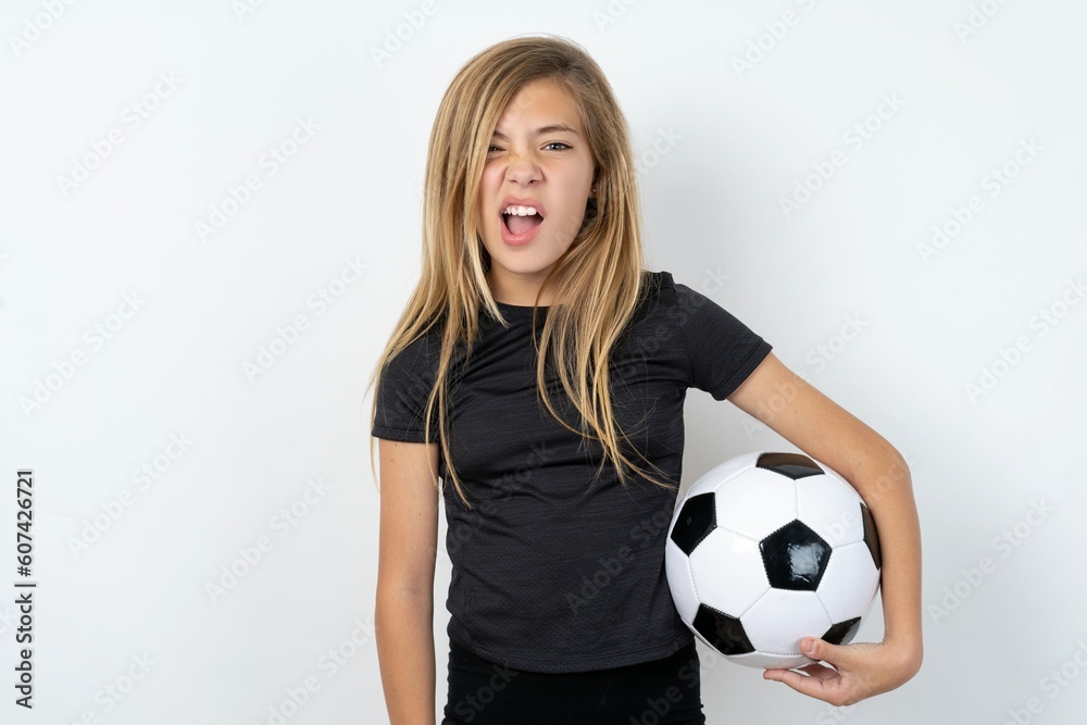 Portrait of dissatisfied beautiful caucasian teen girl wearing sportswear holding a football ball, smirks face, purses lips and looks with annoyance at camera, discontent hearing something unpleasant Portrait of dissatisfied beautiful caucasian teen girl wearing sportswear holding a football ball, smirks face, purses lips and looks with annoyance at camera, discontent hearing something unpleasant