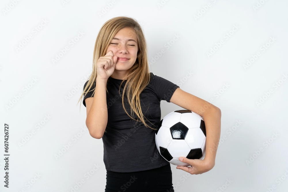 Pleased beautiful caucasian teen girl wearing sportswear holding a football ball over white wall with closed eyes keeps hands near cheeks and smiles tenderly imagines something very pleasant Pleased beautiful caucasian teen girl wearing sportswear holding a football ball over white wall with closed eyes keeps hands near cheeks and smiles tenderly imagines something very pleasant
