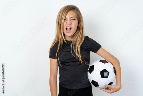 Portrait of dissatisfied beautiful caucasian teen girl wearing sportswear holding a football ball, smirks face, purses lips and looks with annoyance at camera, discontent hearing something unpleasant