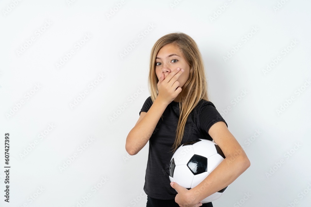 beautiful caucasian teen girl wearing sportswear holding a football ball over white wall covers mouth and looks with wonder at camera, cannot believe unexpected rumors. beautiful caucasian teen girl wearing sportswear holding a football ball over white wall covers mouth and looks with wonder at camera, cannot believe unexpected rumors.