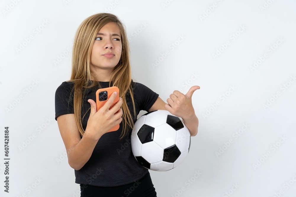 beautiful caucasian teen girl wearing sportswear over white wall points thumb away and shows blank space aside, holds mobile phone for sending text messages. beautiful caucasian teen girl wearing sportswear over white wall points thumb away and shows blank space aside, holds mobile phone for sending text messages.