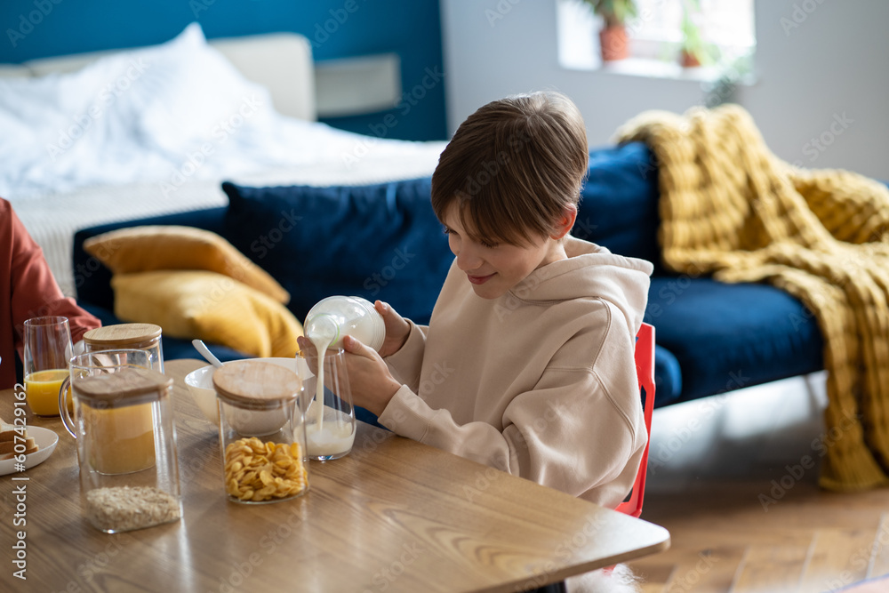 Smiling child schoolboy pouring milk into glass while having healthy breakfast at home, happy kid boy sitting at kitchen table eating cereals. School-aged children and morning meal