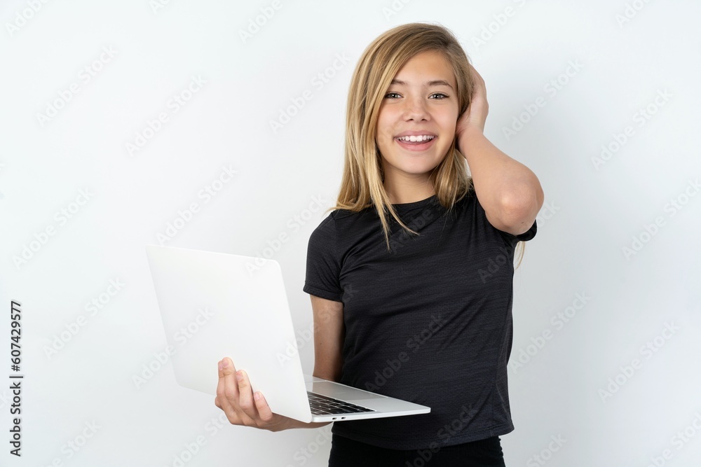 Photo of amazed beautiful caucasian teen girl wearing sportswear over white wall holding white laptop and hand over head. Photo of amazed beautiful caucasian teen girl wearing sportswear over white wall holding white laptop and hand over head.