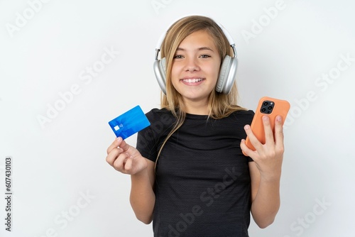 beautiful caucasian teen girl wearing sportswear over white wall wears stereo headphones on ears holds modern mobile phone and credit card