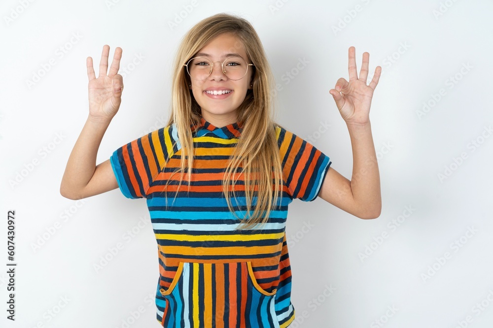 Glad beautiful caucasian teen girl wearing striped T-shirt over white wall shows ok sign with both hands as expresses approval, has cheerful expression, being optimistic. Glad beautiful caucasian teen girl wearing striped T-shirt over white wall shows ok sign with both hands as expresses approval, has cheerful expression, being optimistic.