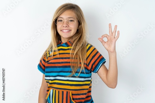 Glad attractive beautiful caucasian teen girl wearing striped T-shirt over white wall shows ok sign with hand as expresses approval, has cheerful expression, being optimistic.