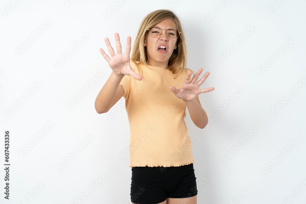 Dissatisfied beautiful caucasian teen girl wearing orange T-shirt over white wall frowns face, has disgusting expression, shows tongue, expresses non compliance, irritated with somebody. Dissatisfied beautiful caucasian teen girl wearing orange T-shirt over white wall frowns face, has disgusting expression, shows tongue, expresses non compliance, irritated with somebody.