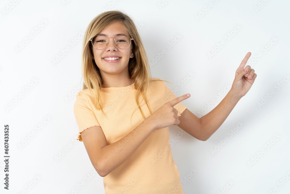 Positive beautiful caucasian teen girl wearing orange T-shirt over white wall with beaming smile pointing with two fingers and looking on empty copy space. Advertisement concept. Positive beautiful caucasian teen girl wearing orange T-shirt over white wall with beaming smile pointing with two fingers and looking on empty copy space. Advertisement concept.