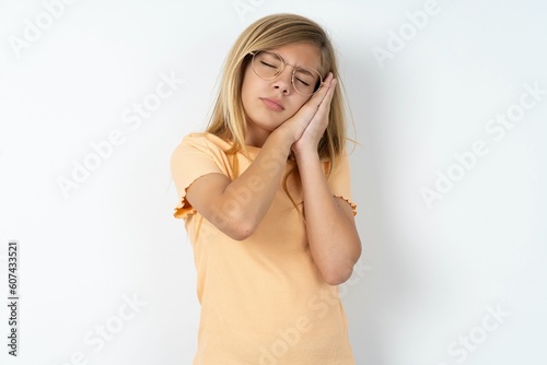 Relax and sleep time. Tired beautiful caucasian teen girl wearing orange T-shirt over white wall with closed eyes leaning on palms making sleeping gesture.