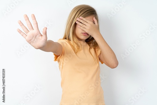 Papier peint beautiful caucasian teen girl wearing orange T-shirt over white wall covers eyes with palm and doing stop gesture, tries to hide