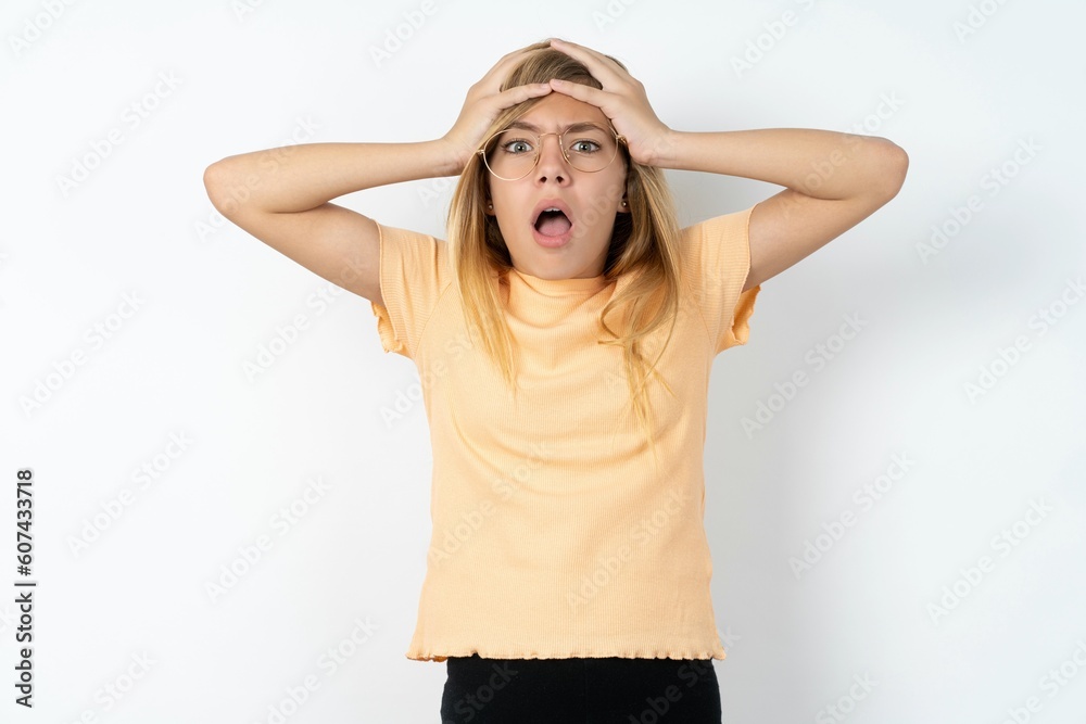 Horrible, stress, shock. Portrait emotional crazy beautiful caucasian teen girl wearing orange T-shirt over white wall clasping head in hands. Emotions, facial expression concept. Horrible, stress, shock. Portrait emotional crazy beautiful caucasian teen girl wearing orange T-shirt over white wall clasping head in hands. Emotions, facial expression concept.
