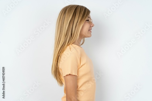 Profile of smiling beautiful caucasian teen girl wearing orange T-shirt over white wall with healthy skin, has contemplative expression, ready to have outdoor walk.