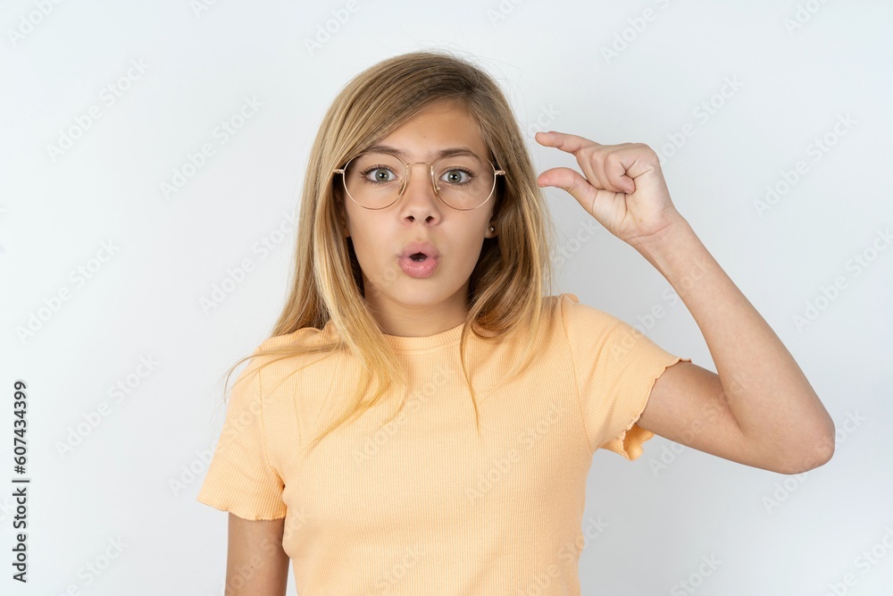 Obraz na plátně Shocked beautiful caucasian teen girl wearing orange T-shirt over white wall shows something little with hands, demonstrates size, opens mouth from surprise