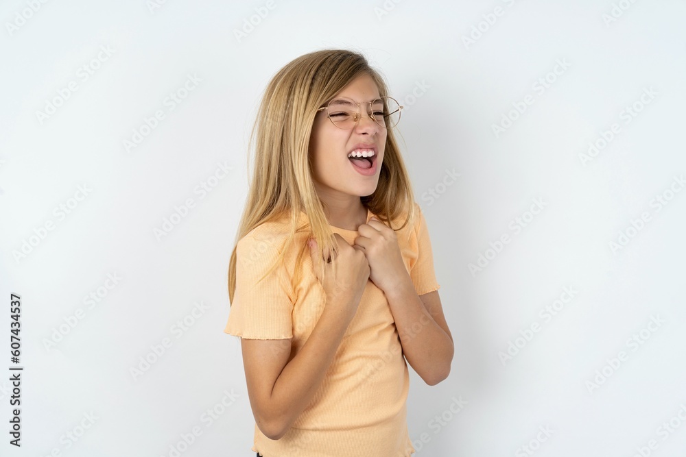 beautiful caucasian teen girl wearing orange T-shirt over white wall excited and glad to achieve victory, clenches fists, screams in excitement with closed eyes,successful person.