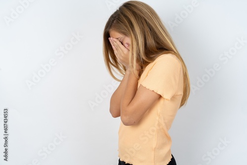 Sad beautiful caucasian teen girl wearing orange T-shirt over white wall covering face with hands and crying.