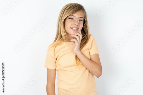 Carefree successful beautiful caucasian teen girl wearing orange T-shirt over white wall touching jawline gazing camera tilting head grinning white teeth delighted. Dental care concept.