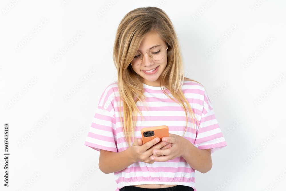 Smiling beautiful caucasian teen girl wearing striped T-shirt over white using cell phone, messaging, being happy to text with friends, looking at smartphone. Modern technologies and communication. Smiling beautiful caucasian teen girl wearing striped T-shirt over white using cell phone, messaging, being happy to text with friends, looking at smartphone. Modern technologies and communication.