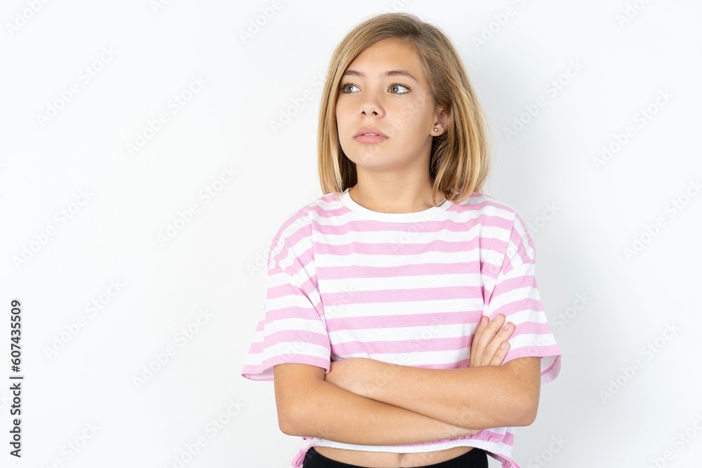 Pleased beautiful caucasian teen girl wearing striped T-shirt over white wall keeps hands crossed over chest looks happily aside Pleased beautiful caucasian teen girl wearing striped T-shirt over white wall keeps hands crossed over chest looks happily aside
