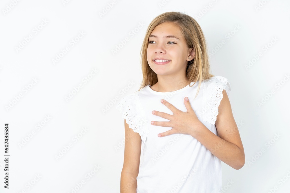 Joyful beautiful caucasian teen girl wearing white T-shirt over white wall expresses positive emotions recalls something funny keeps hand on chest and giggles happily. Joyful beautiful caucasian teen girl wearing white T-shirt over white wall expresses positive emotions recalls something funny keeps hand on chest and giggles happily.
