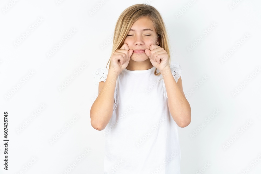 Pleased beautiful caucasian teen girl wearing white T-shirt over white wall with closed eyes keeps hands near cheeks and smiles tenderly imagines something very pleasant Pleased beautiful caucasian teen girl wearing white T-shirt over white wall with closed eyes keeps hands near cheeks and smiles tenderly imagines something very pleasant