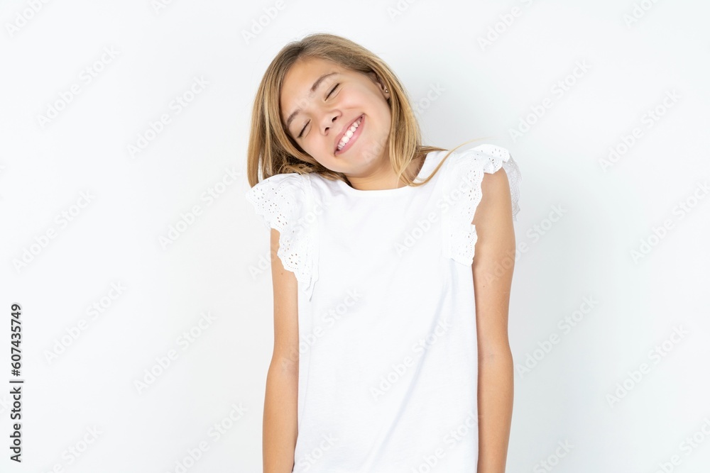 Positive beautiful caucasian teen girl wearing white T-shirt over white wall with overjoyed expression closes eyes and laughs shows white perfect teeth Positive beautiful caucasian teen girl wearing white T-shirt over white wall with overjoyed expression closes eyes and laughs shows white perfect teeth