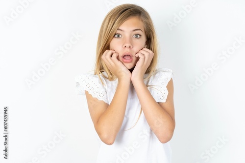 Speechless beautiful caucasian teen girl wearing white T-shirt over white wall keeps hands near opened mouth reacts to shocking news stares wondered at camera