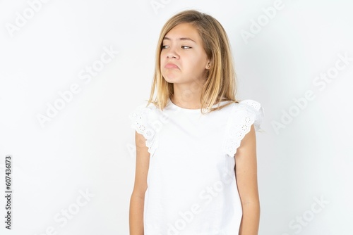 Dissatisfied beautiful caucasian teen girl wearing white T-shirt over white wall purses lips and has unhappy expression looks away stands offended. Depressed frustrated model.