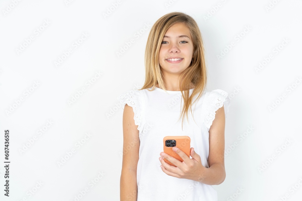 beautiful caucasian teen girl wearing white T-shirt over white wall holds mobile phone in hands and rejoices positive news, uses modern cellular beautiful caucasian teen girl wearing white T-shirt over white wall holds mobile phone in hands and rejoices positive news, uses modern cellular
