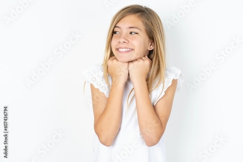 beautiful caucasian teen girl wearing white T-shirt over white wall holds hands under chin, glad to hear heartwarming words from stranger