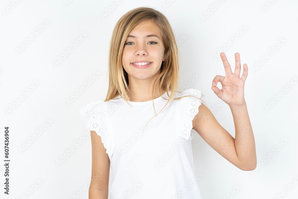 beautiful caucasian teen girl wearing white T-shirt over white wall hold hand arm okey symbol toothy approve advising novelty news beautiful caucasian teen girl wearing white T-shirt over white wall hold hand arm okey symbol toothy approve advising novelty news
