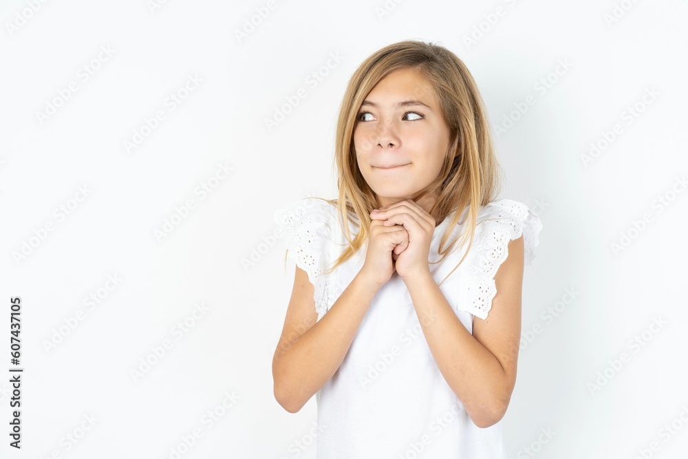 Curious beautiful caucasian teen girl wearing white T-shirt over white wall keeps hands under chin bites lips and looks with interest aside. Curious beautiful caucasian teen girl wearing white T-shirt over white wall keeps hands under chin bites lips and looks with interest aside.