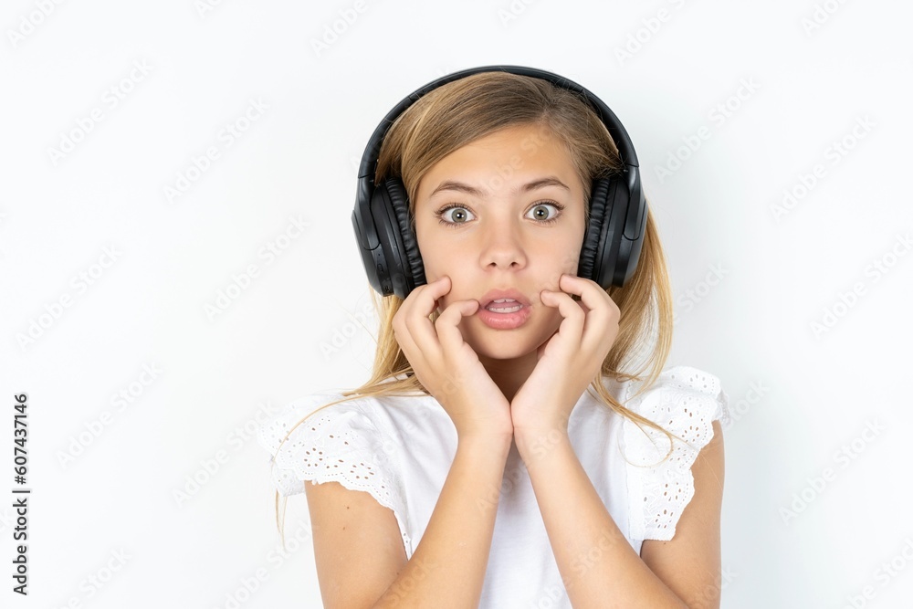 Shocked beautiful caucasian teen girl wearing white T-shirt over white wall stares fearful at camera keeps mouth widely opened wears wireless stereo headphones on ears Shocked beautiful caucasian teen girl wearing white T-shirt over white wall stares fearful at camera keeps mouth widely opened wears wireless stereo headphones on ears