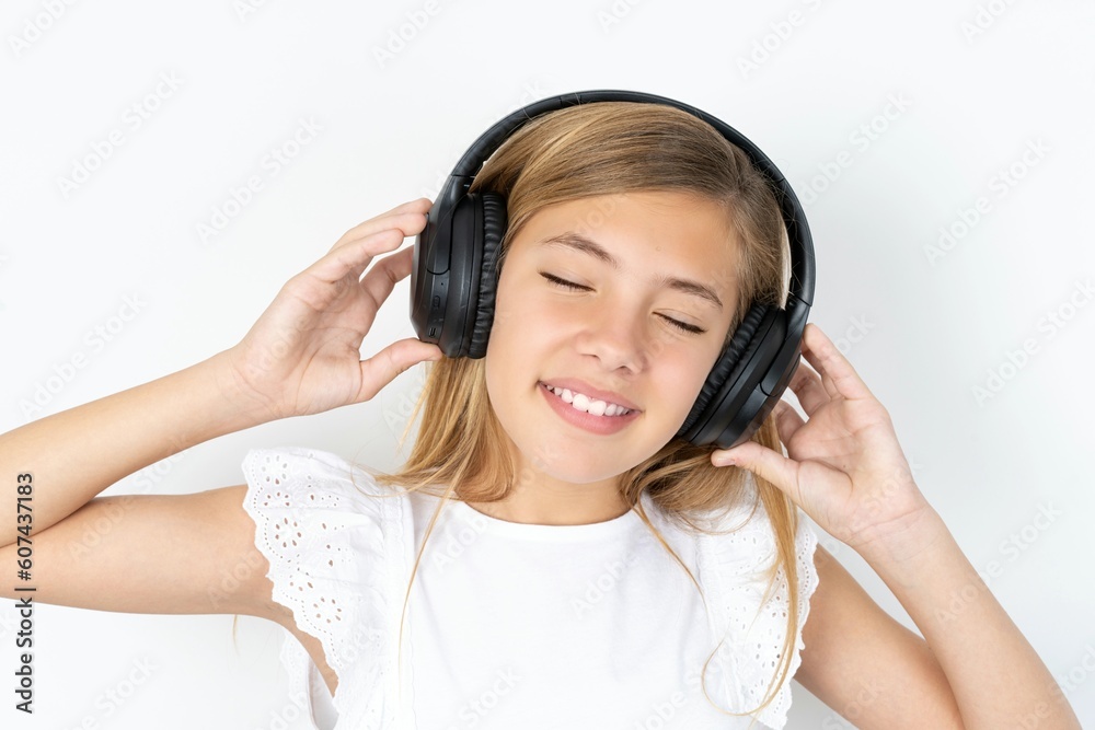 beautiful caucasian teen girl wearing white T-shirt over white wall smiles broadly feels very glad listens favourite music track via wireless headphones closes eyes. beautiful caucasian teen girl wearing white T-shirt over white wall smiles broadly feels very glad listens favourite music track via wireless headphones closes eyes.