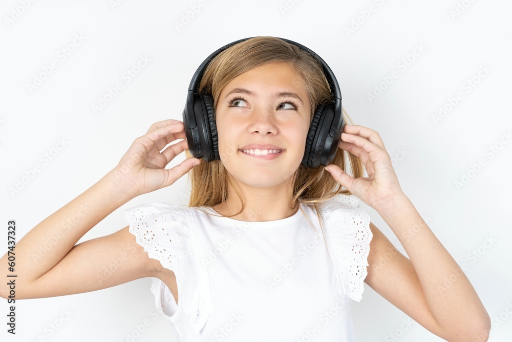 Joyful beautiful caucasian teen girl wearing white T-shirt over white wall sings song keeps hand near mouth as if microphone listens favorite playlist via headphones Joyful beautiful caucasian teen girl wearing white T-shirt over white wall sings song keeps hand near mouth as if microphone listens favorite playlist via headphones