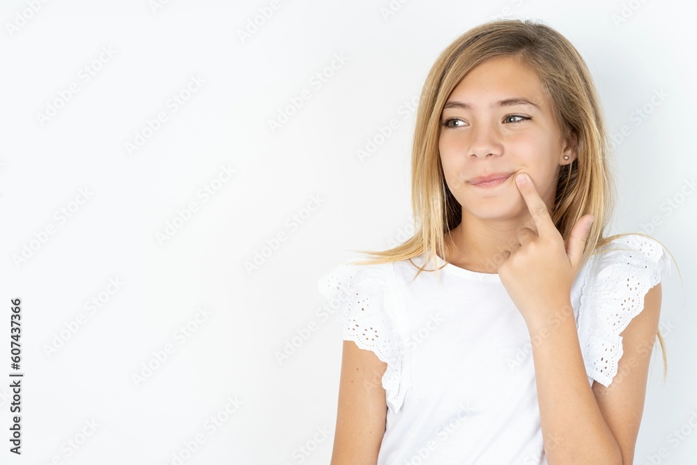 Lovely dreamy beautiful caucasian teen girl wearing white T-shirt over white wall keeps finger near lips looks aside copy space. Lovely dreamy beautiful caucasian teen girl wearing white T-shirt over white wall keeps finger near lips looks aside copy space.