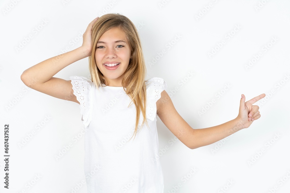 Surprised beautiful caucasian teen girl wearing white T-shirt over white wall pointing at empty space holding hand on head