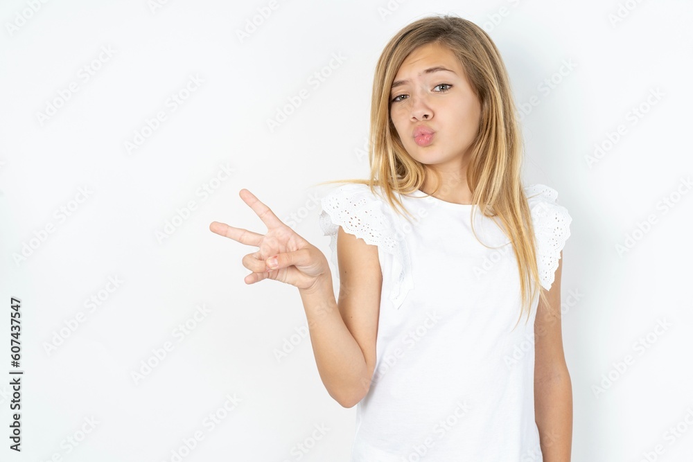 Obraz premium beautiful caucasian teen girl wearing white T-shirt over white wall makes peace gesture keeps lips folded shows v sign. Body language concept