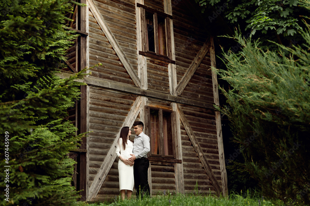 Fototapeta premium beautiful pregnant couple on the background of a wooden ecological house in the forest. a man hugs a pregnant woman.